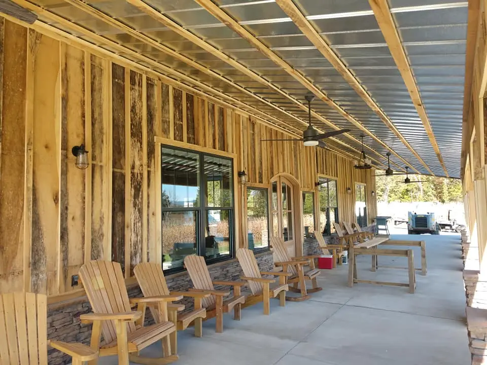 Long covered porch with a metal roof, ceiling fans, rough-sawn board siding, stone wainscot, and wooden Adirondack chairs